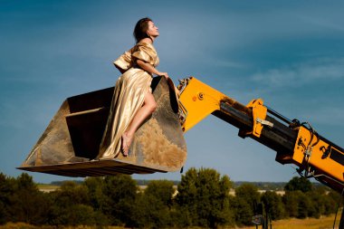 Captivating woman in an elegant golden dress poses gracefully in a construction vehicle bucket against a backdrop of a clear blue sky and expansive field landscape, creating a striking contrast.