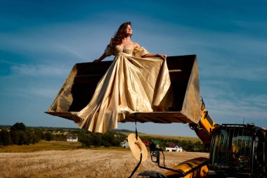Captivating Woman in Elegant Gold Dress Posing on Tractor Loader Bucket in Golden Field, a Striking Contrast of Glamour and Rural Landscape Under a Bright Blue Sky, Straw Hat Hanging from Above.