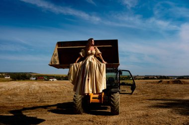 Captivating image of a gorgeous woman in a luxurious gown gracefully seated within an excavator bucket set against a stunning blue sky, creating a striking contrast and unique outdoor scene, fashion meeting industry.