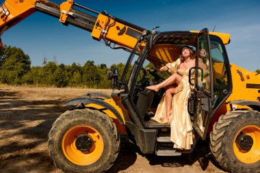 Glamorous woman in a golden dress exudes confidence, sitting barefoot in a vibrant yellow telescopic handler on a sunlit construction site, creating a striking juxtaposition of elegance and industry.