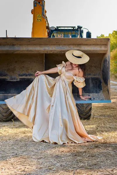 A stunning woman in a gold dress strikes a pose on construction equipment in a rural setting, the juxtaposition of elegance and industry creates striking scene.