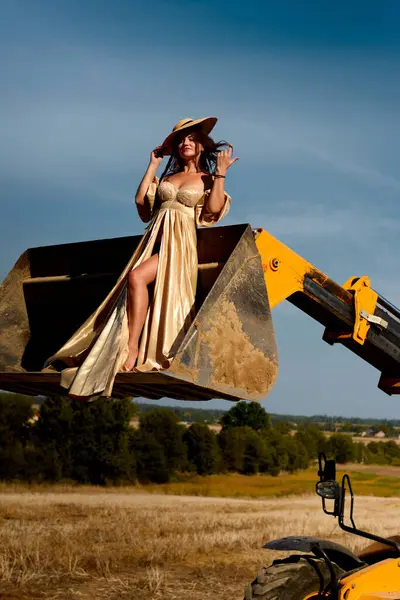 Elegant woman in a golden gown and straw hat striking a pose on a yellow excavator bucket in a harvested field, blending fashion and industry against a bright sky.
