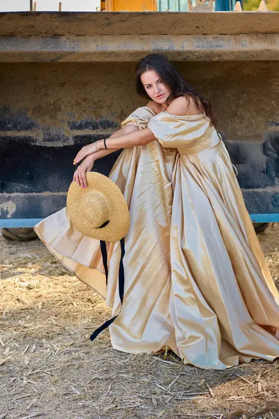 A poised brunette in a luxurious gold gown strikes a pose against heavy machinery, the straw hat with a black ribbon adding a touch of vintage elegance, capturing the essence of timeless beauty amidst industrial elements.