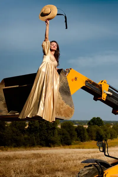 Elegant woman in a luxurious gold dress posing in tractor bucket, holding straw hat against the field and blue sky on summer sunny day, outdoor fashion photoshoot, concept of village life.
