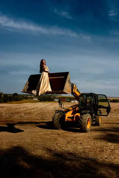 Elegant woman in a long golden dress gracefully poses inside tractor's bucket in golden wheat field during sunset, embodying surreal blend of beauty, agriculture and fashion.