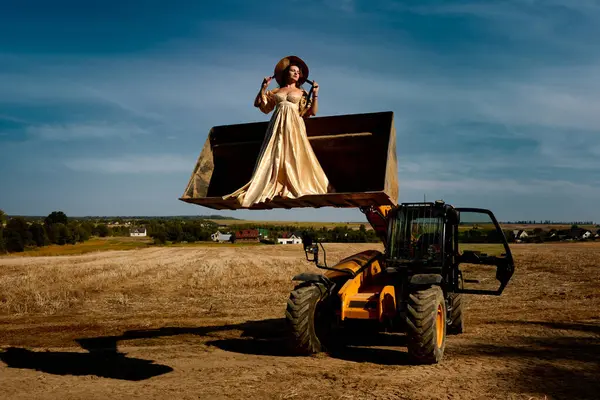 Beautiful woman in elegant golden dress and straw hat stands in the tractor bucket in rural harvested field with houses on background under bright blue sky, unique fashion concept.