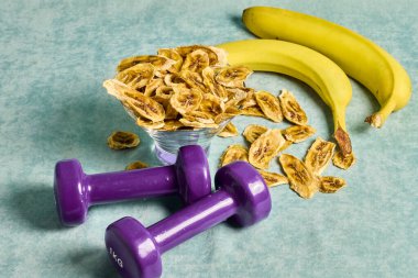 Healthy lifestyle balance featuring dumbbells, fresh bananas for energy, and a bowl of dried banana chips, symbolizing fitness and healthy eating choices, set on a textured table.