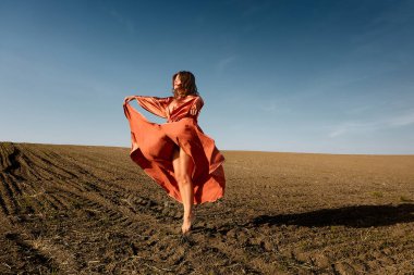 A barefoot woman in a copper dress dances freely in a plowed field with the blue sky above, showcasing movement, beauty, and the harmony of fashion and nature against a rustic backdrop.