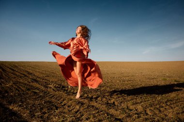 A beautiful woman in an elegant red dress dances freely in a sunlit field under a clear blue sky, expressing joy and liberation, with her flowing gown and barefoot movement creating a serene and graceful scene, while capturing the essence of nature