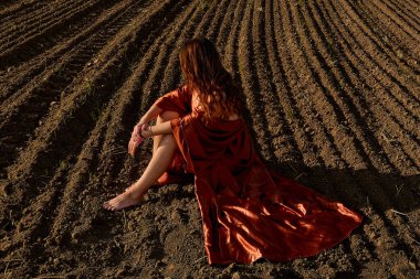 A captivating image shows a thoughtful woman in a copper gown, resting in a freshly tilled field during the sunset, creating a scene of serenity and natural beauty under the golden hour lighting.
