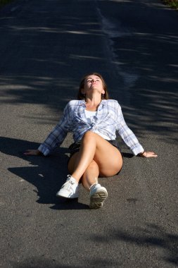 A young woman in casual attire, sitting cross-legged on a country road in sunshine, enjoys a moment of peace with arms spread wide, embracing the warm summer day and the open road, looking peaceful.
