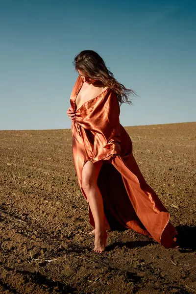 Elegant woman in a flowing orange dress walks barefoot on ploughed ground under a serene sky, radiating freedom, beauty, and connection with the earth, embodying natural grace.