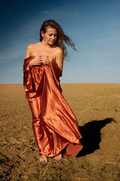 A serene moment captured as a young woman, wrapped in copper-colored fabric, stands barefoot in a harvested field, lost in thought with a downward gaze on a bright, sunny day.
