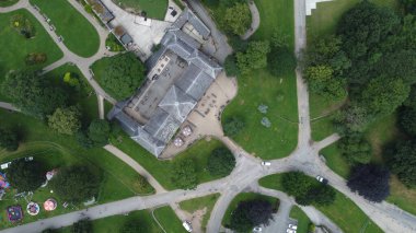 An aerial perspective shows a sprawling park featuring a substantial building, winding pathways, and vibrant green trees. Visitors enjoy outdoor activities on a sunny day.