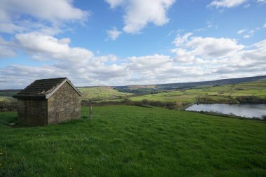 Parlak yeşil tarlalar ve bulutlu bir gökyüzünün altındaki uzak tepelerle çevrili huzurlu bir göle bakan çimenli bir tepenin üzerinde kırsal taştan bir kulübe duruyor..