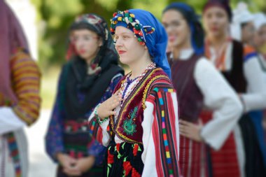 Svistov, Bulgaristan - 07 / 27 / 2024: Bulgaristan 'ın Gradeznitsa kentinden geleneksel Pomak kostümü. Kadın şalvari pantolon, nakışlı gömlek, yelek ve ponponlu mavi başörtüsü giyiyor. Geleneksel kıyafetlerle görünen diğer kadınlar, 