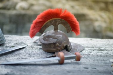 Bir kılıcın yanına yıpranmış taş bir yüzeye yerleştirilmiş parlak kırmızı dumanlı Roma tarzı bir kask. Tarihi içerik, antik Roma veya askeri canlandırmalar için ideal.. 