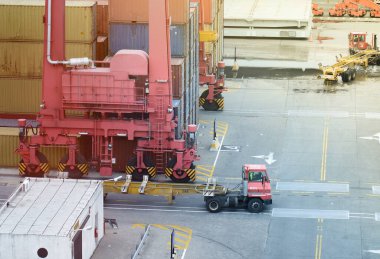 Kırmızı vinçleri olan yoğun bir konteynır portu ve konteyner iskelesini hareket ettiren bir terminal traktörü, kargo taşımacılığı ve nakliye lojistiğinin endüstriyel sürecini gösteriyor..