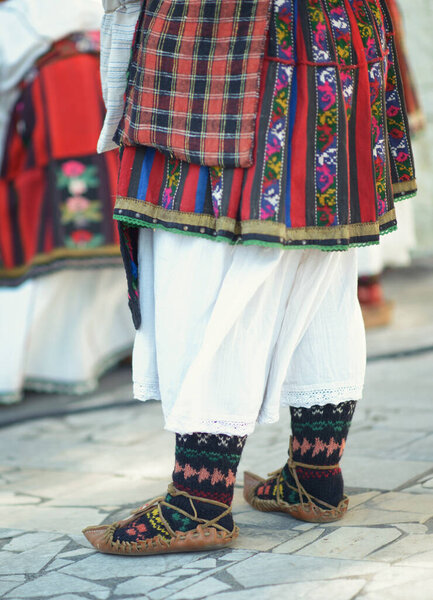 Intricate layers of Bulgarian attire featuring a handmade crossbody bag, colorful embroidered skirt, white underskirt, patterned socks, and traditional leather shoes.