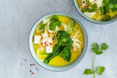 Pirinç erişteli vegan körili laksa, brokoli ve tofu mavi kaselerde, gri arka plan, üst manzara.