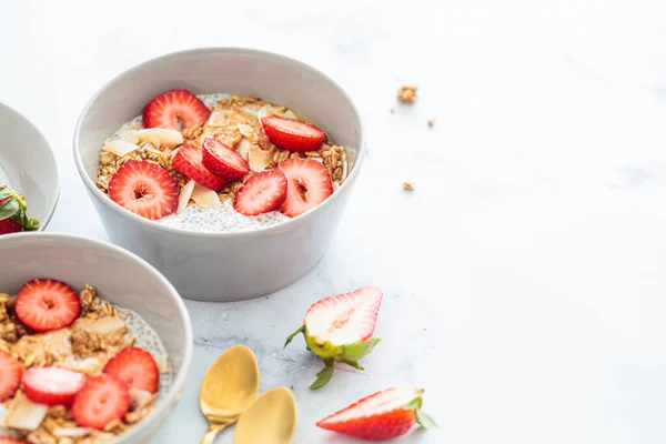 Chia pudding with homemade coconut granola, peanut butter and strawberries in gray bowl, marble background. Healthy plant based diet, detox, summer recipe.