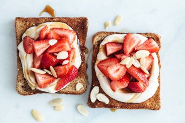 Flat lay of breakfast toasts with ricotta, strawberries and almonds on a white marble background.