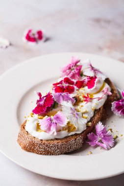 Kahvaltı krem peynirli tost pembe mutfak çiçekleri tabakta, pembe mermer arka planda.