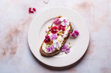 Kahvaltı krem peynirli tost pembe mutfak çiçekleri tabakta, pembe mermer arka planda, üst manzara.