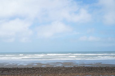 Sörf ve Bulutların Altındaki Çakıl Zeminli Sakin Sahil: Yumuşak dalgaların farklı bulut örtüleri olan bir gökyüzünün altında kıyıda buluştuğu çakıl taşları ve kumsalın huzurlu bir görüntüsü.