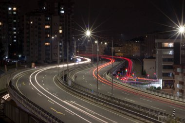 A dynamic night long-exposure photograph showcasing the vibrant light trails of cars on a curving city highway. Bright starburst streetlights illuminate the scene, with apartment buildings and urban infrastructure forming the backdrop, capturing.