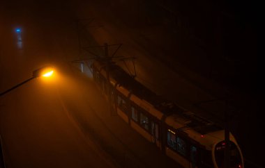 Orange glowing street lamp on a very foggy dark night with tram in background. Night scene with street lighting illuminating dense atmospheric fog and railroad tracks