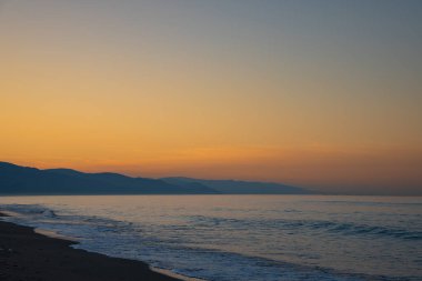 Kumsalda gün batımı ve dağlar. Ufku olan akşam plajı manzarası. Siluet, doğa, sükunet, manzara harikası ve kutsal güzellik konsepti