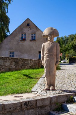 Büyük bir şapka takan bir adamın uzun taş heykeli kaldırım taşından bir patikanın yanında konumlandırılmıştır. Arkasında berrak mavi gökyüzünün altında yıpranmış bir bina var, sakin bir köy atmosferi yaratıyor..