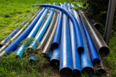 Lacivert ve siyah borulardan oluşan bir koleksiyon yeşil çimlerin üzerinde yer alıyor. Bu da potansiyel bir inşaat ya da bahçe projesini işaret ediyor. Yerleşim huzurlu ve doğal, biraz fazla gelişmiş bitki örtüsüyle..