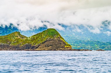 Puslu kıyı manzarası, engebeli tepeler yeşilliklerle kaplı, dalgalar kayalık kıyılara çarpıyor, ve arka planda orman dağları var. Kaliteli fotoğraf. Uvita Puntarenas Eyaleti Kosta