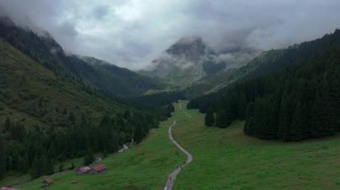 Sinema insansız hava aracı, çam ormanları ve küçük kulübeleriyle sisli bir dağ vadisinde yükseliyor. Kamera yukarı doğru hareket ediyor ve tepelerin etrafındaki dramatik bulutları ortaya çıkarıyor. Huzurlu ve gizemli dağlık arazi.