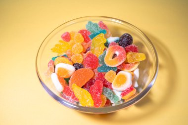 Sarı arka planda çeşitli canlı, şeker kaplı şekerlerle dolu cam bir kasenin genel manzarası..