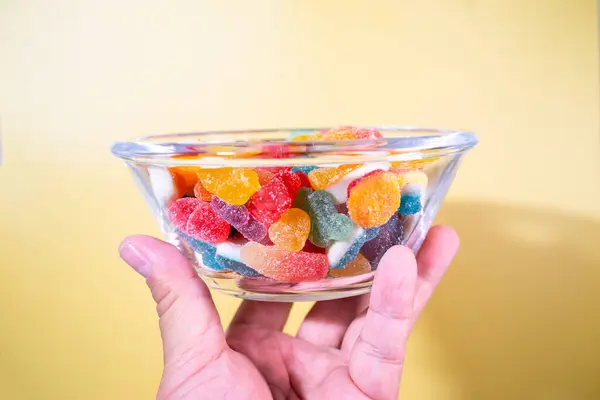 Sarı arka planda çeşitli canlı, şeker kaplı şekerlerle dolu cam bir kasenin genel manzarası..