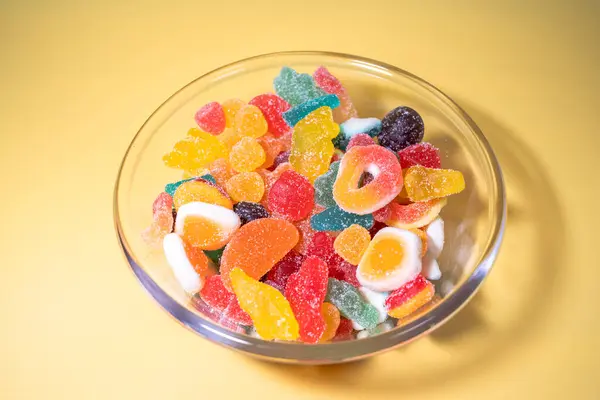 Sarı arka planda çeşitli canlı, şeker kaplı şekerlerle dolu cam bir kasenin genel manzarası..