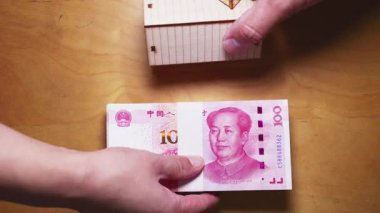 İki el Çin Yuan banknotlarını küçük ahşap bir model evle değiş tokuş ediyor. Tahta bir masa üzerindeki emlak yatırımını ve emlak işlemlerini simgeliyor.