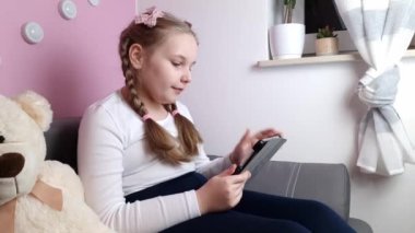 Little girl sitting on sofa and waving her hand at screen of tablet. Modern technology, communication, childhood concepts