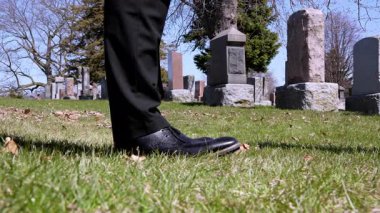 Cinematic insert shot of man's feet in cemetery dropping wilted flower bouquet, with tombstones in the background.