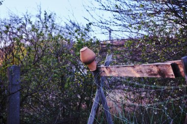 A weathered clay pot sits upside down on a rustic wooden fence post in a natural, overgrown garden setting. The image evokes a sense of rural life, tradition, and simplicity.