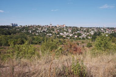 Scenic panoramic view of a city residential area nestled on a green hill, surrounded by lush trees under a bright blue sky. Captures the harmony between urban development and nature on a clear, sunny day.