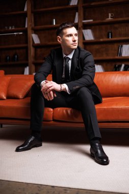 Elegant young man in black suit posing in a modern studio interior with bookshelves and leather sofa, stylish business portrai