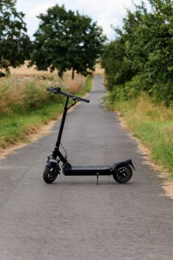 A sleek black electric scooter standing on an empty rural asphalt road surrounded by summer greenery and golden dry grass. The image conveys eco-friendly transportation, freedom, and a peaceful countryside vibe.