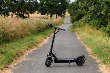 A sleek black electric scooter standing on an empty rural asphalt road surrounded by summer greenery and golden dry grass. The image conveys eco-friendly transportation, freedom, and a peaceful countryside vibe.