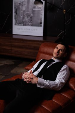 Elegant young man in black suit posing in a modern studio interior with bookshelves and leather sofa, stylish business portrait