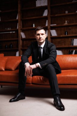 Elegant young man in black suit posing in a modern studio interior with bookshelves and leather sofa, stylish business portrait
