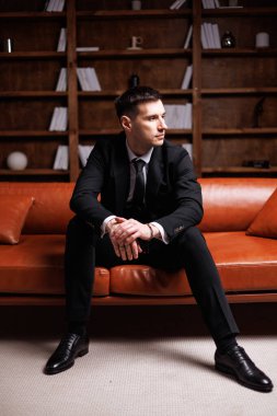Elegant young man in black suit posing in a modern studio interior with bookshelves and leather sofa, stylish business portrait